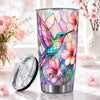 A clear insulated tumbler with a stainless steel base and a colorful stained-glass-style design of a hummingbird and pink flowers. The lid is placed beside it on a marble surface, with pink flowers and soft-focus flowers in vases in the background.